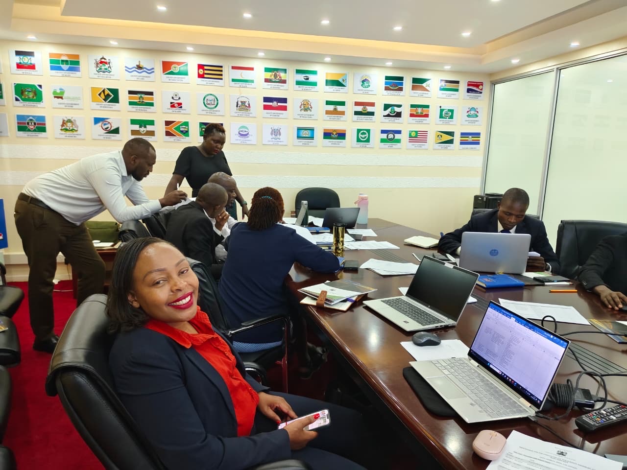 Participant smiling in a county government boardroom lined with Kenyan county flags during a working session